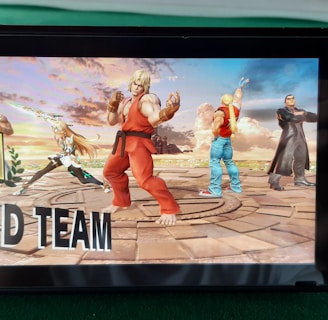 A handheld gaming console displays a screenshot of a fighting game featuring cartoonish characters in dynamic poses. The characters are standing in a virtual arena setting with a sky and cloud backdrop. The console is placed on a green surface.